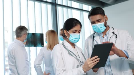 Mixed-Races-Couple,-Man-And-Woman,-Doctors-In-Medical-Masks-Talking-And-Using-Tablet-Device