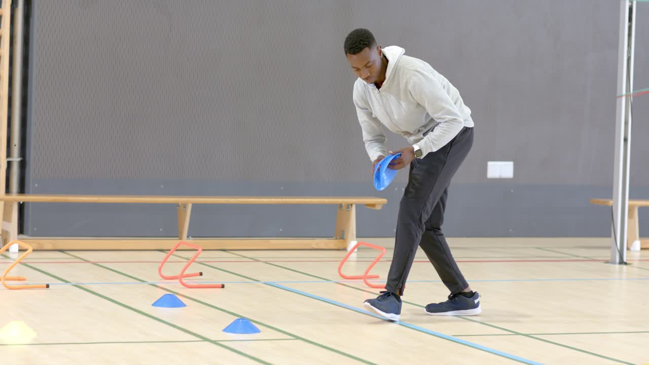Afroamerikansk mandlig træner går ind i sportshal og placerer skiver ved hække til en volleyballøvelse