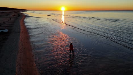 una mujer está caminando a lo largo de una playa de arena, en el borde del océano, una fuerte puesta de sol se puede ver en la distancia, que deleita con sus colores brillantes
