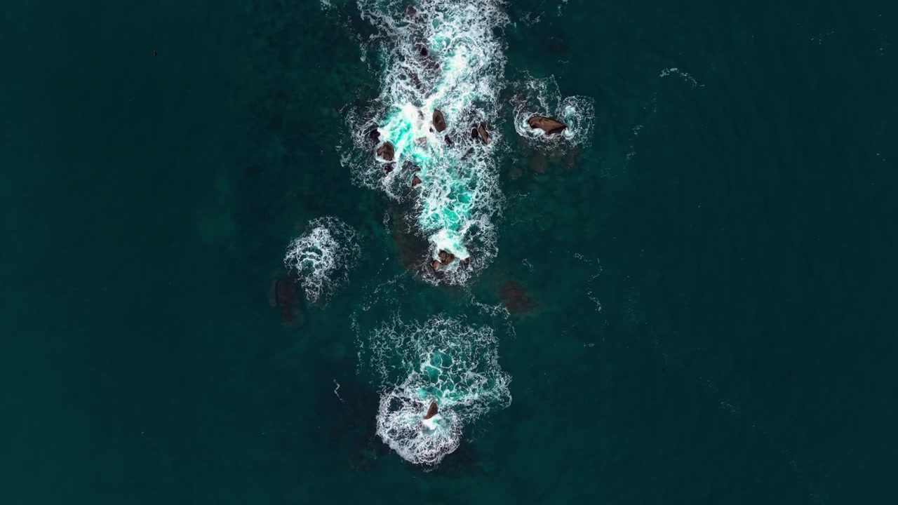 Aerial view of white surf swirling around submerged dark stones in dark teal ocean waters. Overhead perspective of isolated reef rocks in deep sea.