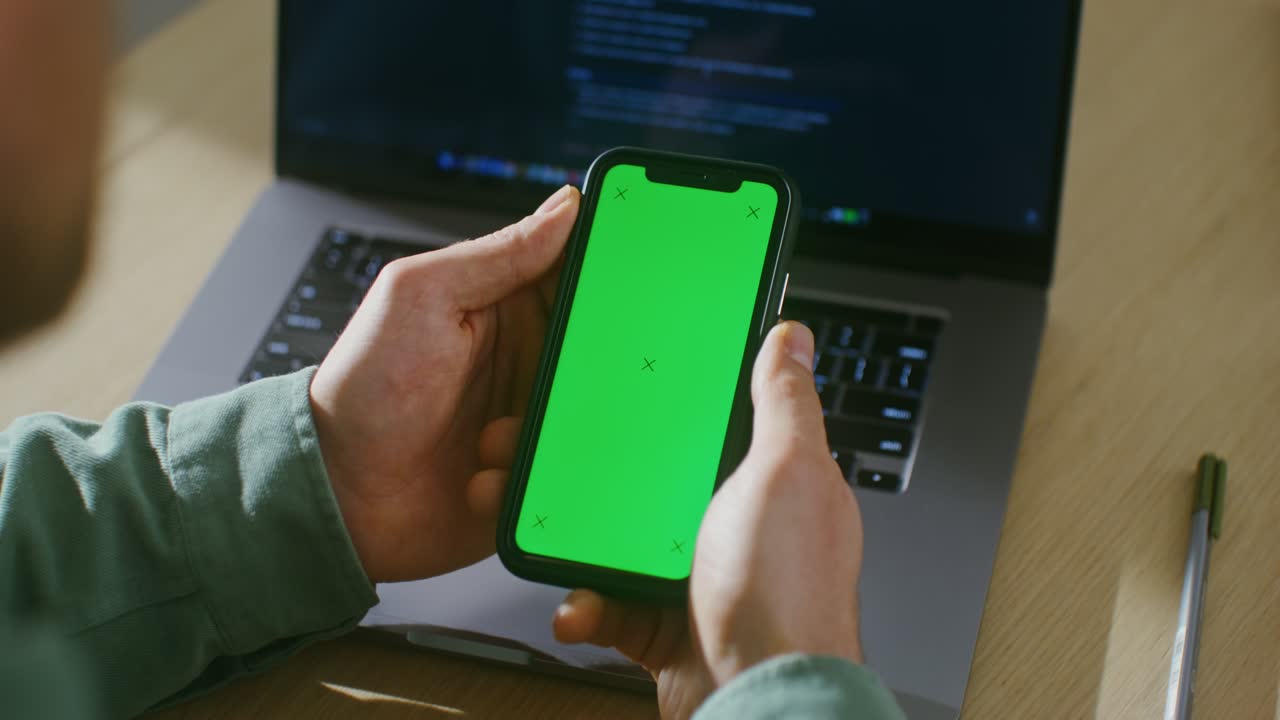 persona que sostiene un teléfono inteligente con pantalla verde frente a una computadora portátil