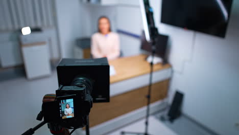 blog content creation in studio. woman sits at desk in the room looking at professional camera. blurred backdrop.