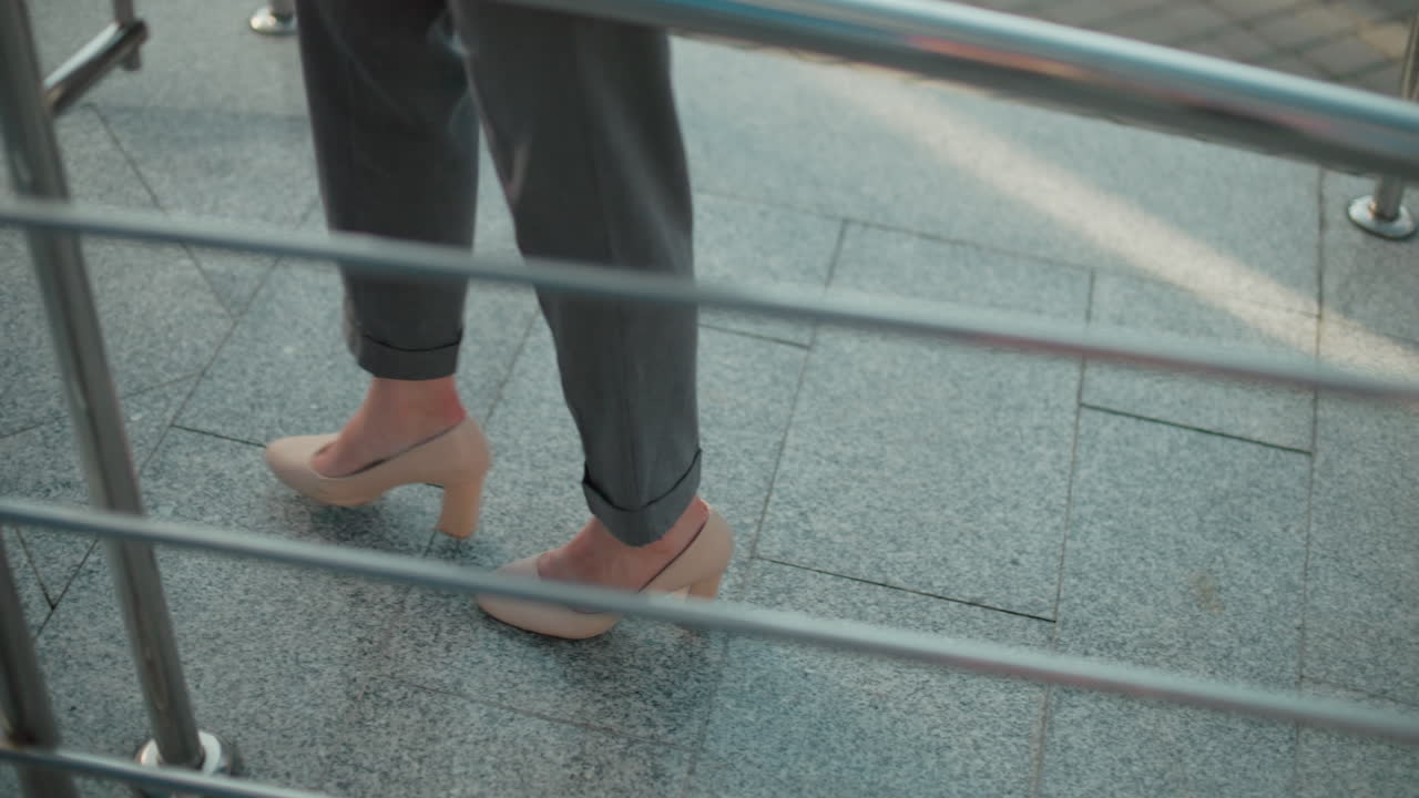 Leg view of woman in nude color heels walking confidently by iron railing on tiled pavement in outdoor urban setting, showing elegant stride and professional attire with sunlight casting soft shadows