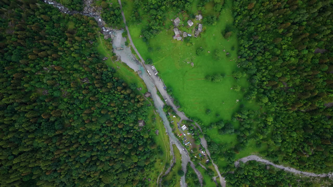 Drone shot in full top-down view orbiting and descending over a river with a road alongside it, surrounded by alpine forest and scattered houses in the Swiss Alps