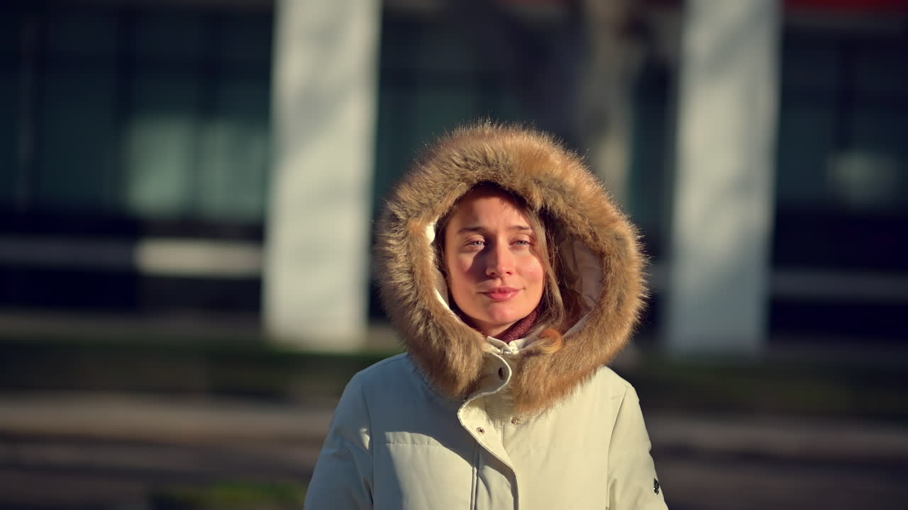 A woman stands outside wearing a cozy winter coat with a fur-lined hood. She smiles contentedly as she enjoys the warmth of the sun in a city park with modern buildings in the background