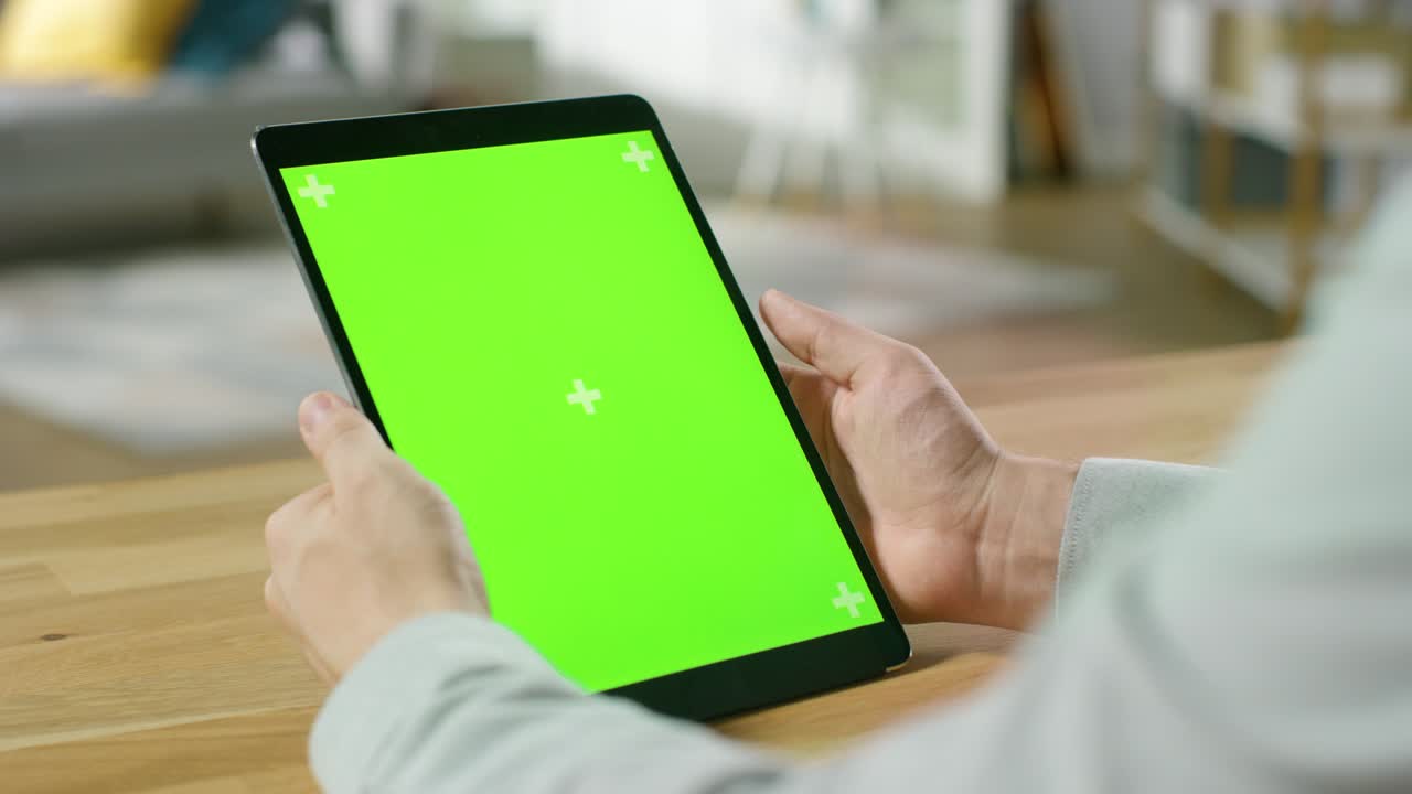 Mand bruger håndbevægelser på en digital tabletcomputer med grøn mock-up skærm i portrættilstand, mens han sidder ved sit skrivebord. I baggrunden er der en hyggelig stue.