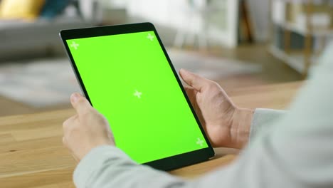 hombre usando gestos de la mano en pantalla de maqueta verde computadora tableta digital en modo retrato mientras está sentado en su escritorio. en el fondo acogedor salón.