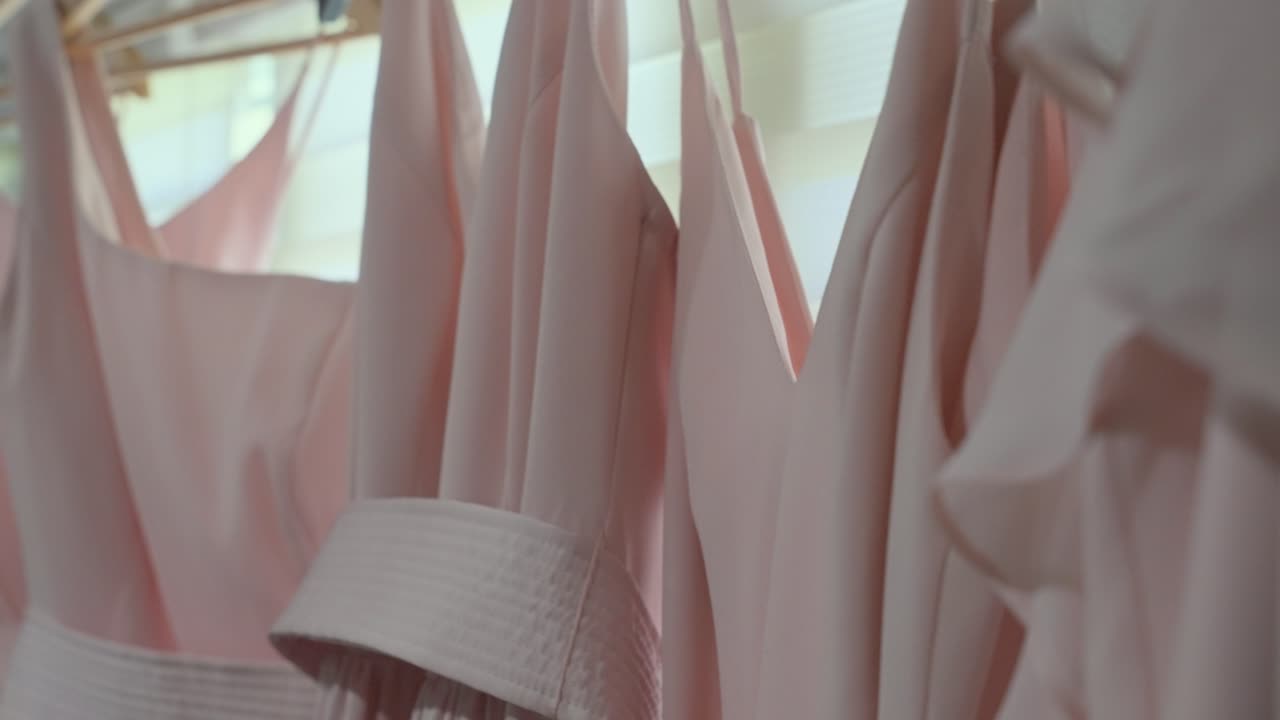 Una fluida ripresa con tilt verticale che mostra una fila di abiti da damigella rosa cipria abbinati, con scollature e dettagli in tessuto diversi, appesi in preparazione di una cerimonia nuziale