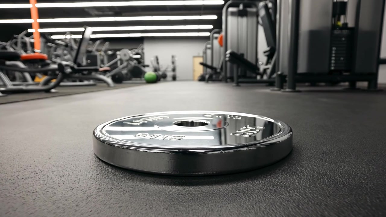 Placa de peso cromada brilhante repousa em um piso de borracha escuro dentro de uma academia moderna e limpa, com máquinas de força desfocadas, luzes de teto lineares brilhantes e espaço de treino aberto criando um ambiente de fitness focado.