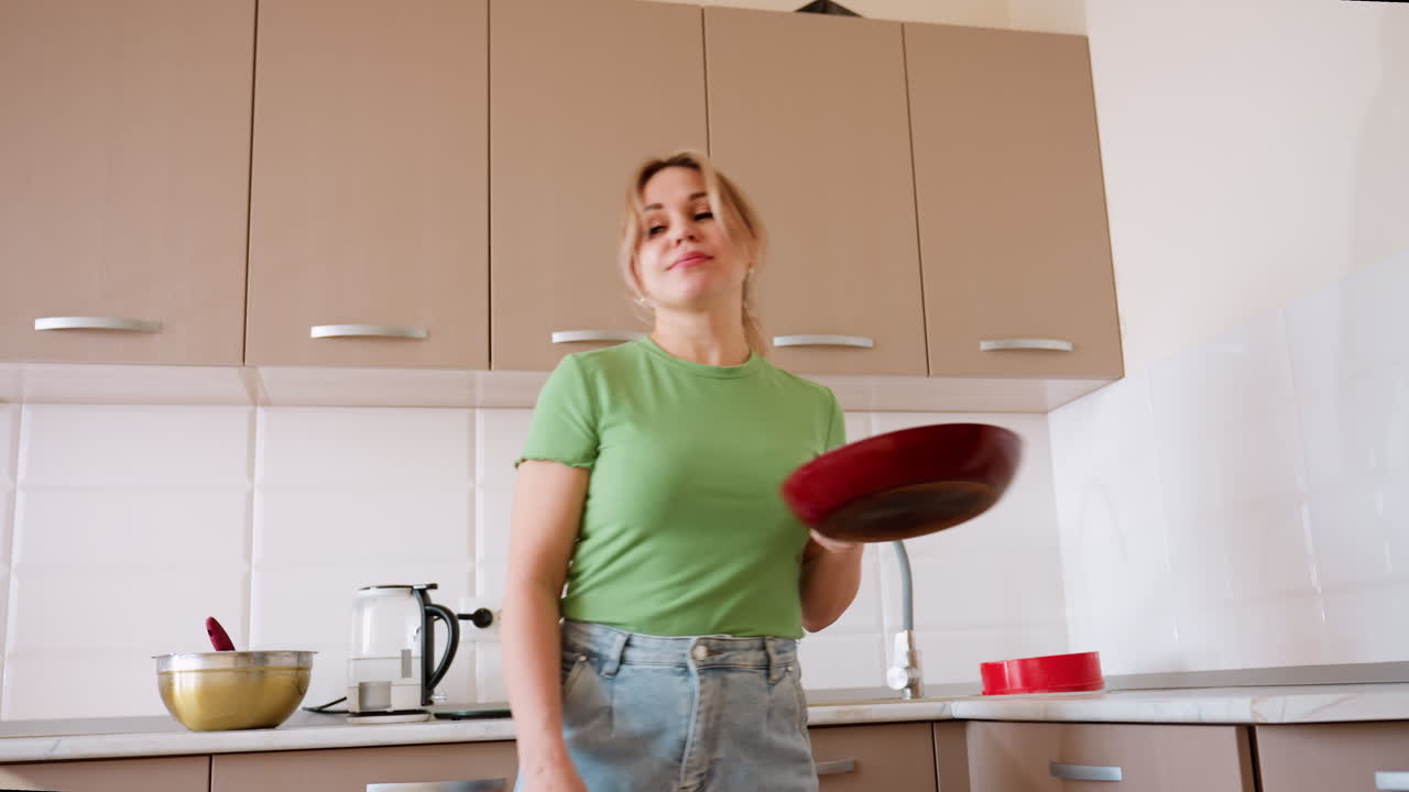 Donna con maglietta verde e jeans prende una padella dal cassetto della cucina durante la preparazione del cibo in una cucina luminosa con bollitore, ciotola, teglia rossa, lavandino e mobili in legno sullo sfondo