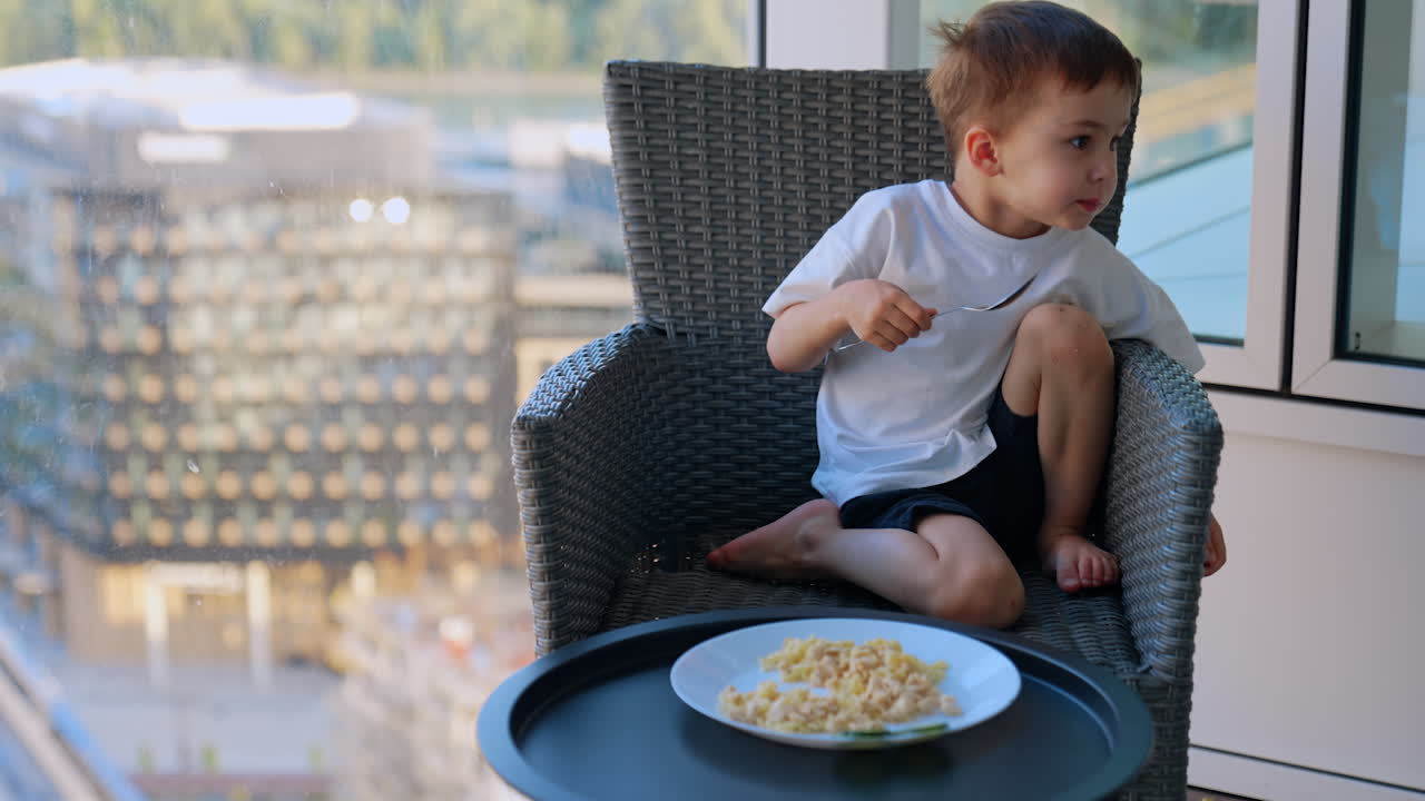 Bambino che pranza sul balcone. Un giovane ragazzo è seduto su una sedia da balcone, mangiando pasta da un piatto con vista su un paesaggio urbano.