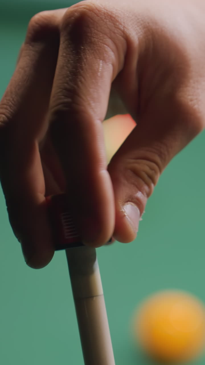 Close-up da mão de um homem passando giz em um taco de sinuca antes de uma tacada em uma mesa de bilhar verde. Foco na precisão e na técnica, com bolas de bilhar borradas ao fundo. Movimento suave, jogo profissional