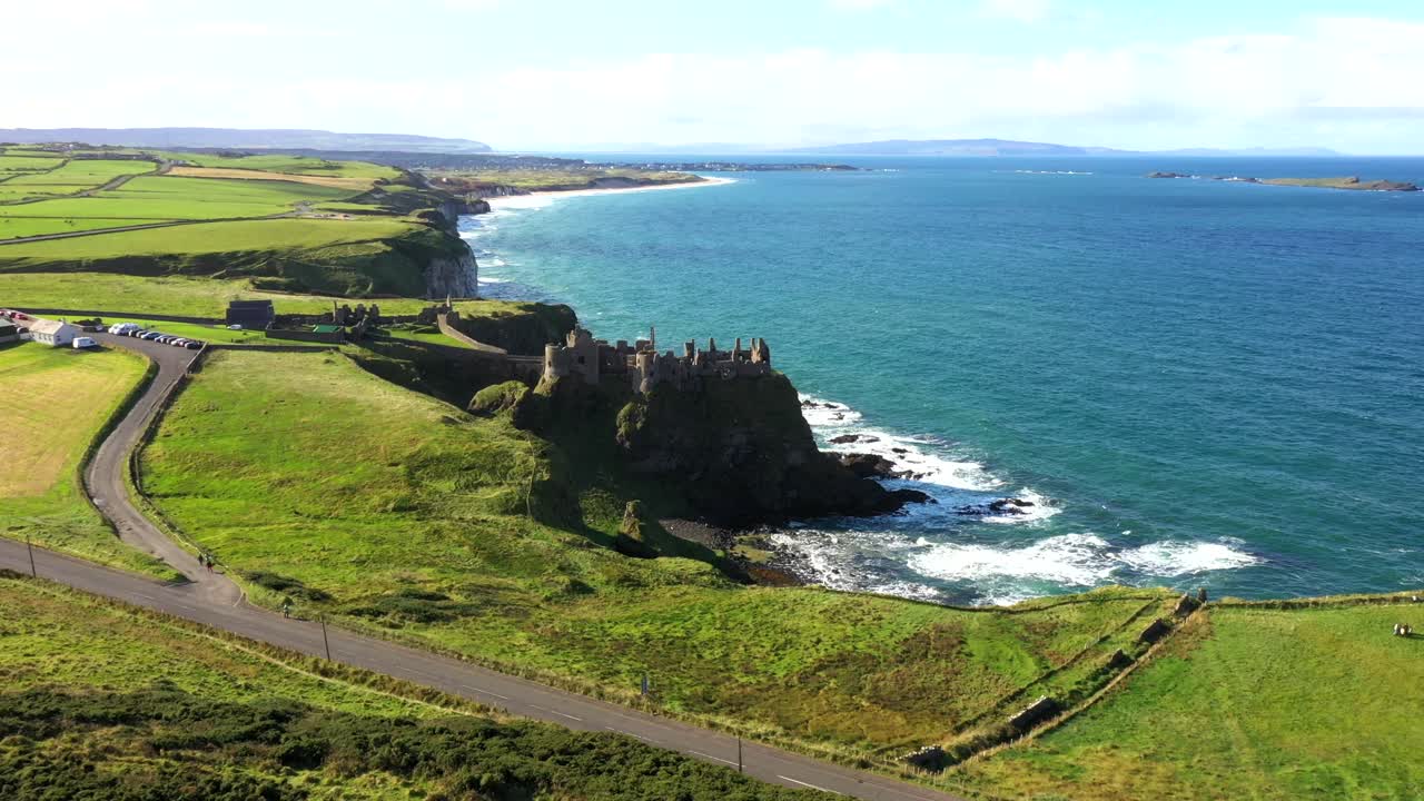 High drone flyover of green clifftop land extending toward sea horizon in Northern Ireland, Dunluce castle centered on rocky outcrop