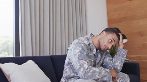 thoughtful biracial male soldier in uniform sitting on sofa in living room, slow motion