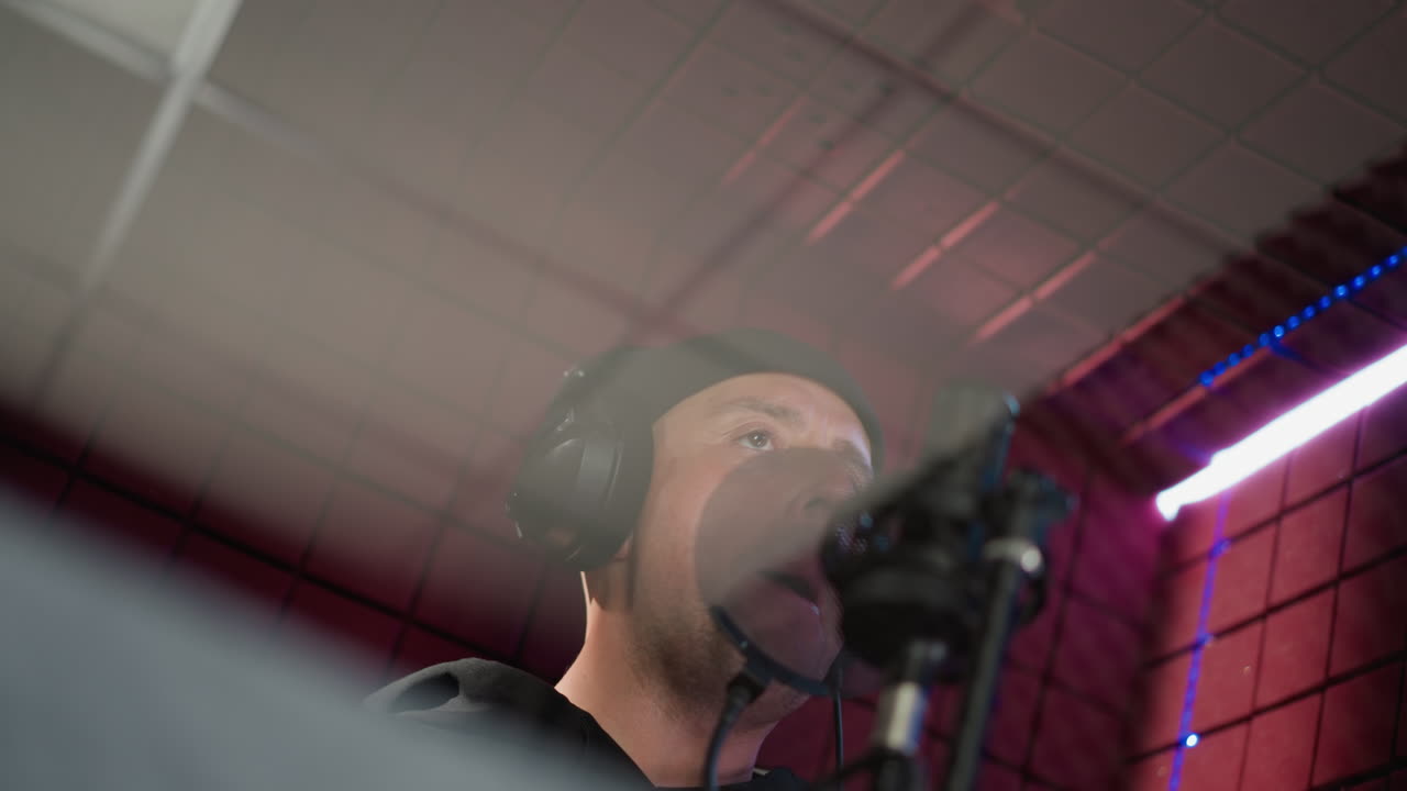 A man dressed in black is wearing headphones and standing in front of a microphone in a red soundproofed room. The image captures the moment before he starts singing, with expressive hand movements