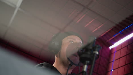 a man dressed in black is wearing headphones and standing in front of a microphone in a red soundproofed room. the image captures the moment before he starts singing, with expressive hand movements