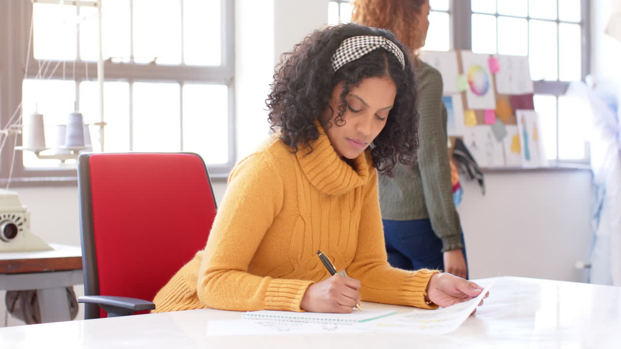 Happy biracial female fashion designer drawing a design in studio, slow motion