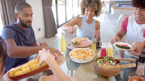 Diverse friends eating burgers, fries, and salad, enjoying meal together at home