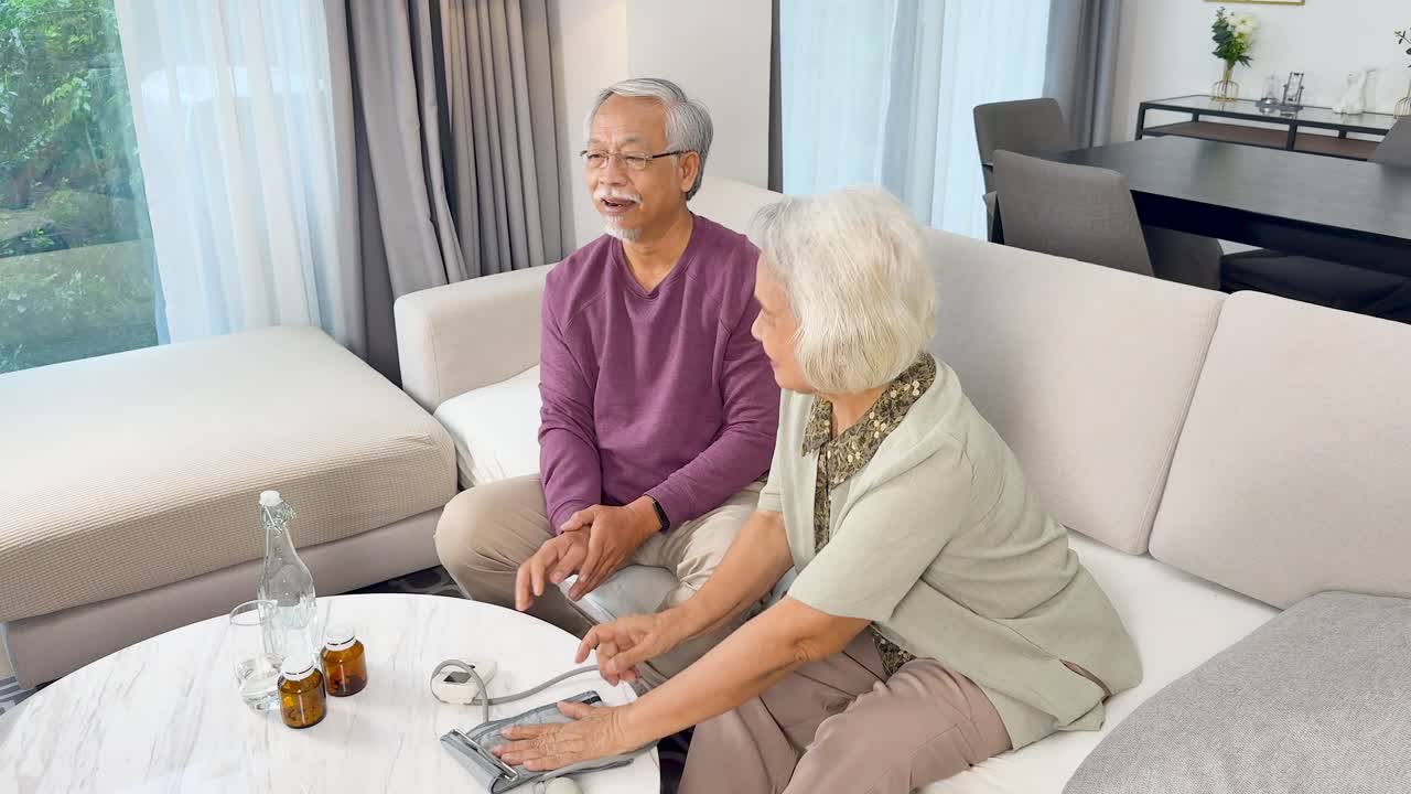 Elderly couple uses digital blood pressure monitor together in bright, cozy living room setting