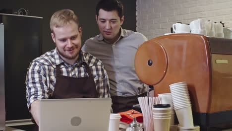 coffee shop owner and manager discussing data on laptop near espresso machine, manager laughing, slow motion medium shot