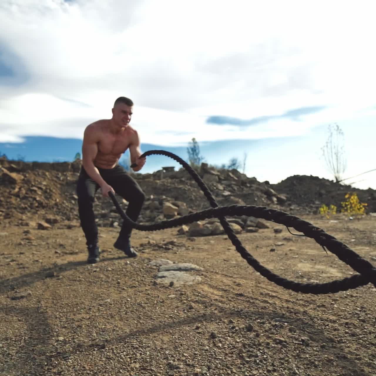 Athletic male performs battle ropes exercise at sunlight. Handsome shirtless man working with battle ropes during an outdoor fitness workout in slow motion.