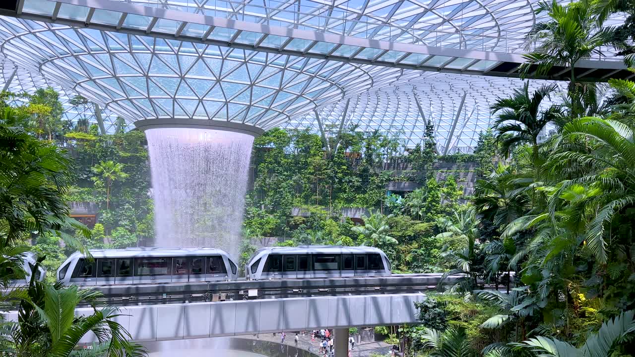 An automated train glides past a large indoor waterfall surrounded by dense greenery under a glass dome, with bright natural lighting and a steady camera angle