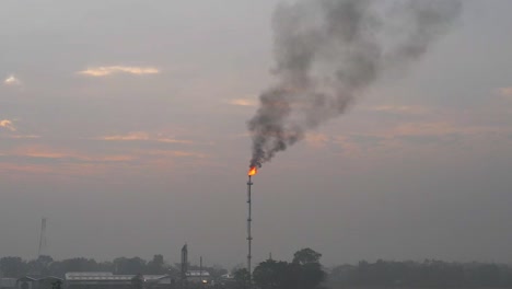 Petrochemical-refinery-flaming-smoking-flare-stack-burning-above-misty-agricultural-farmland-at-sunrise