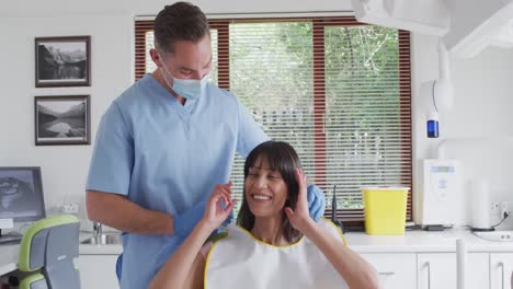 Dentista-Masculino-Caucásico-Con-Mascarilla-Preparando-A-Una-Paciente-Sonriente-En-Una-Clínica-Dental-Moderna