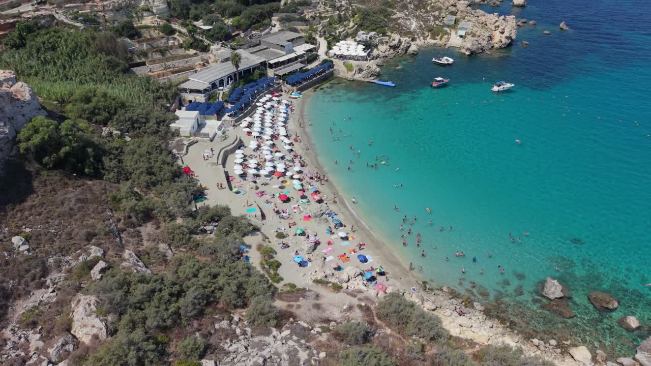 An aerial video of a sandy beach in Malta filled with colorful umbrellas and sunbeds, where tourists enjoy the clear waters. People are swimming, relaxing on the shore, and boats are anchored nearby