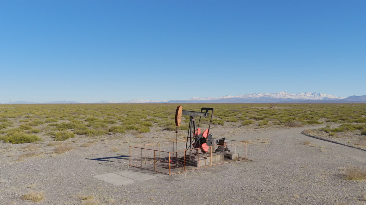 Drone captures oil pumpjack operating in dry steppe near Mendoza, Argentina, with snowy Andes peaks in background under clear blue sky, wide and slow pull away shot