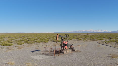 drone captures oil pumpjack operating in dry steppe near mendoza, argentina, with snowy andes peaks in background under clear blue sky, wide and slow pull away shot