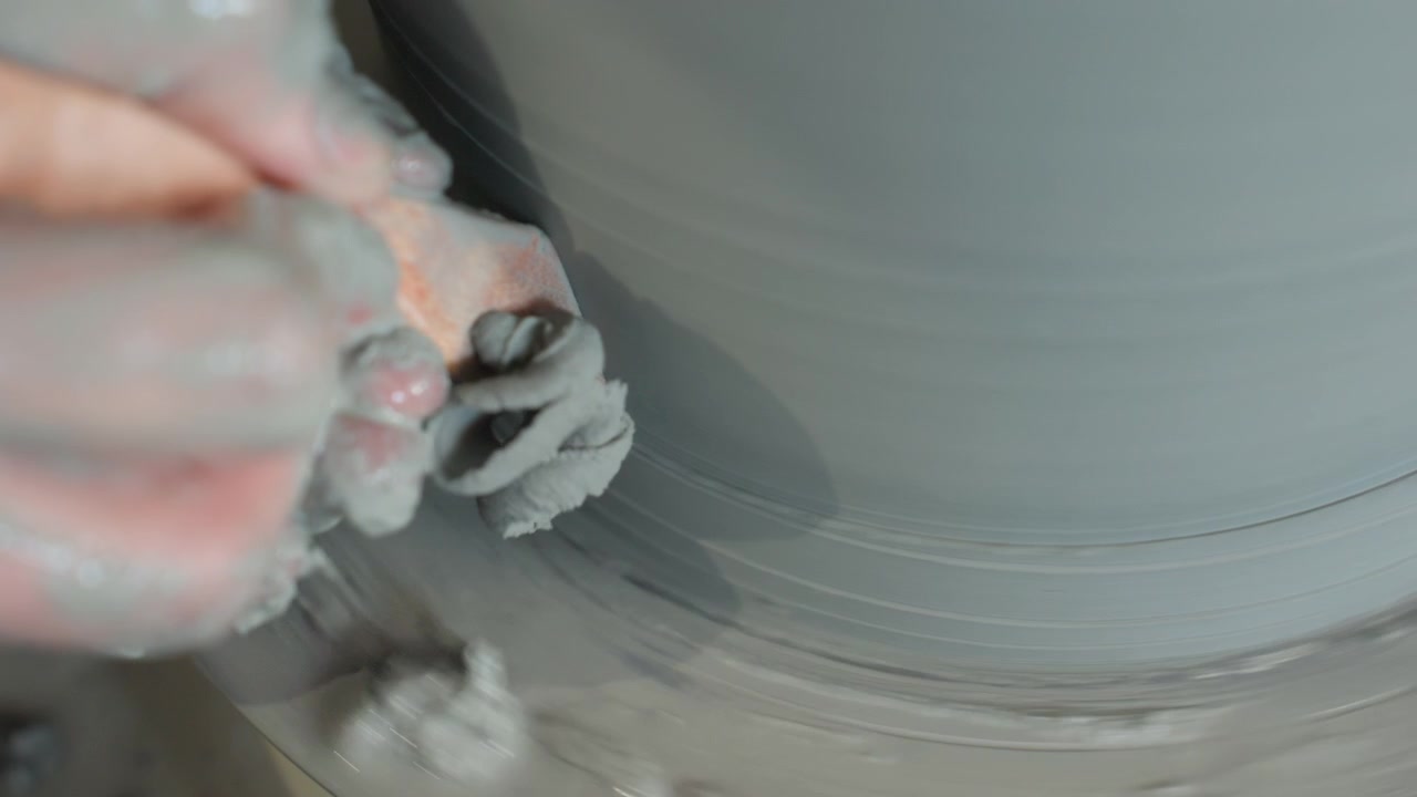 Hands meticulously carving thin clay shavings carefully. Detailed view of ceramic shaping on spinning wheel. Focus on delicate pottery rim trimming using paddle tool during steady studio practice