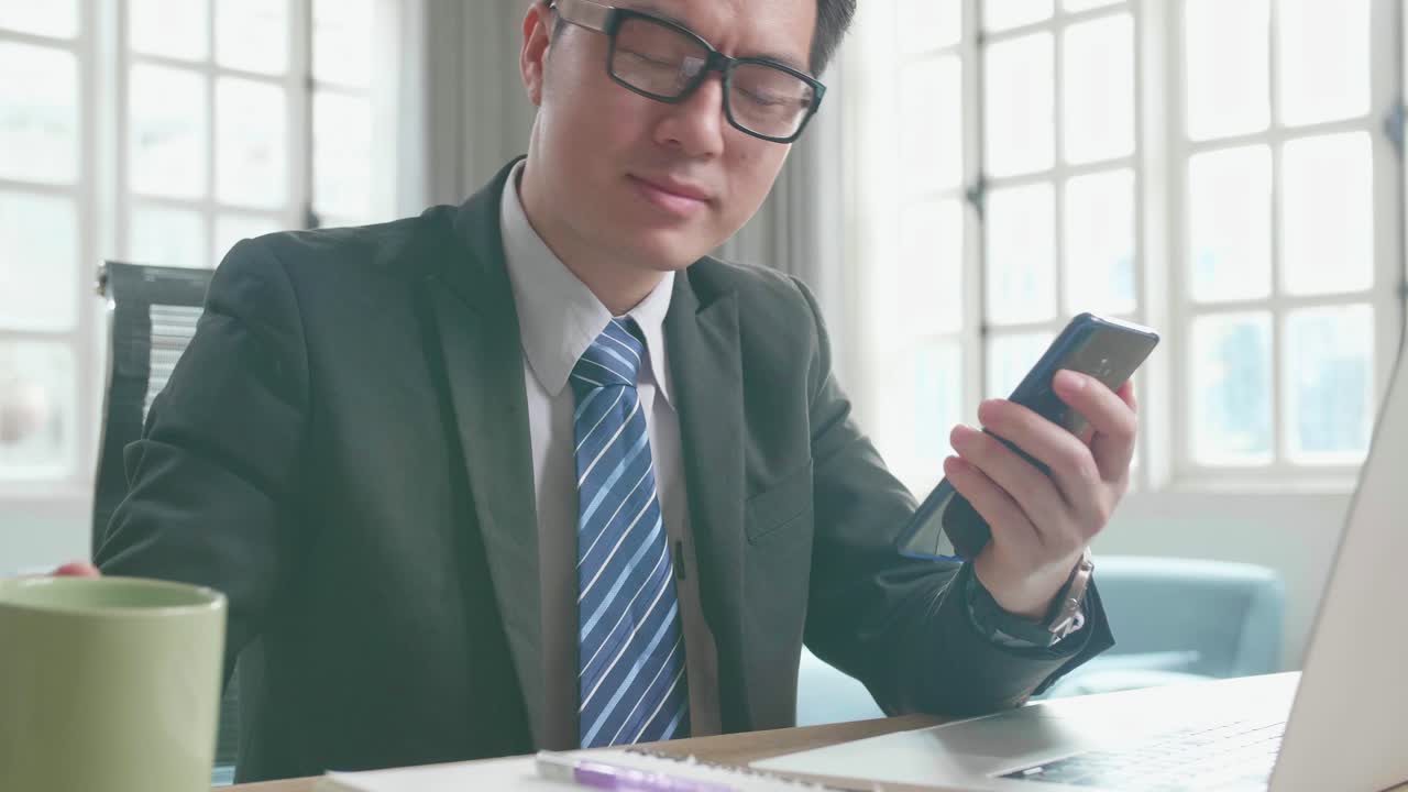 Asian Businessman With Glasses Wearing Business Suit Using Mobile Phone And Drinking Coffee While Using The Computer For Working At Home.