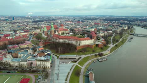 Establishing-dolly-moving-forward-aerial-over-Wawel-Royal-Castle-by-the-river-Vistula-in-the-city-of-Krakow,-Poland,-with-the-city-center-in-the-background-at-sunset