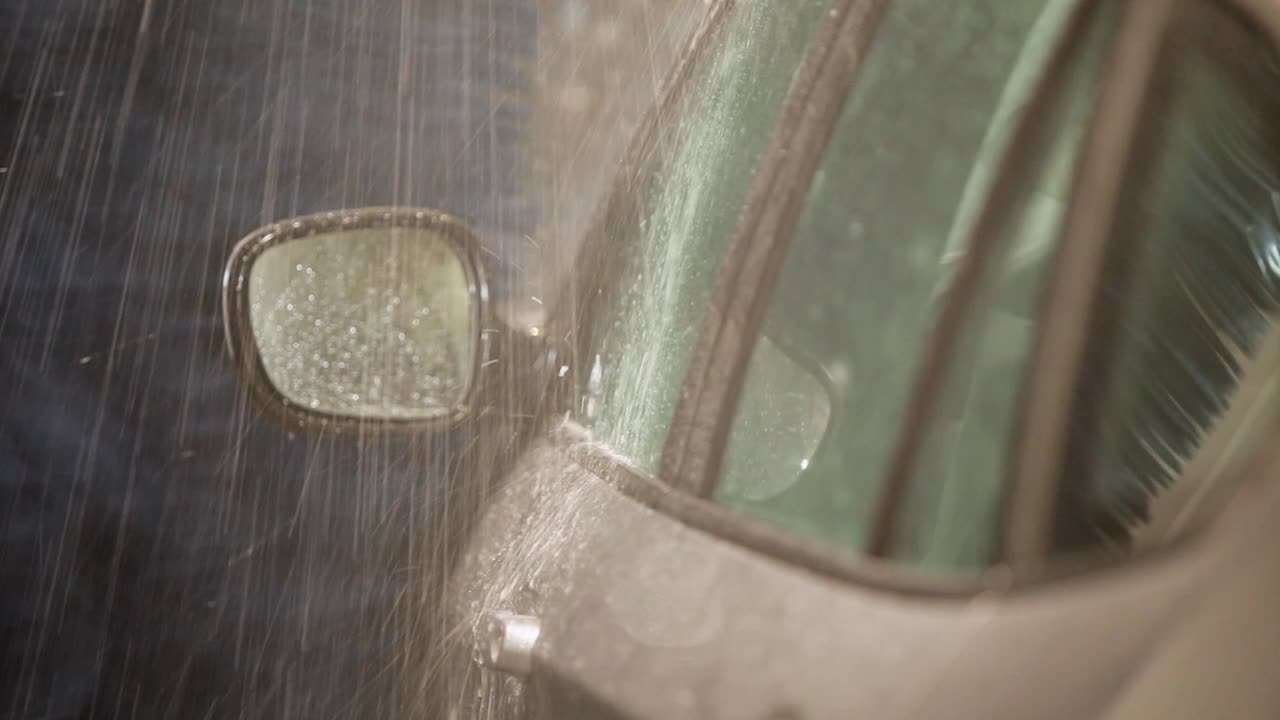 In the grasp of a handheld camera, a close-up of a white car's round side mirror with a silver frame going into an automated car wash