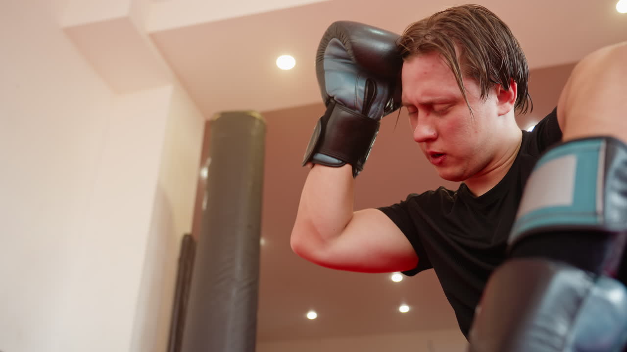 Male fighter resting after sparring in gym, wearing black sportswear, face showing fatigue and determination while breathing heavily, looking focused and thoughtful under training lights