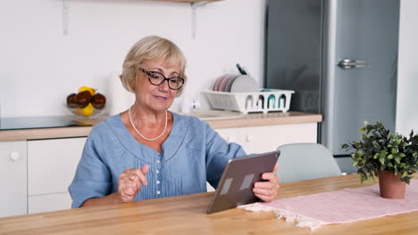 happy senior woman sitting at table in kitchen greeting and talking on video call using a digital tablet