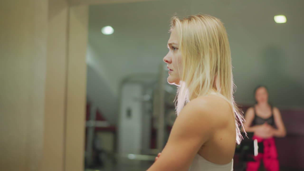 atleta solitaria che si riflette nello specchio della palestra, donna bionda a braccia incrociate e sguardo pensieroso, luce brillante dello studio dietro, profilo muscoloso, concentrazione pre-allenamento e determinazione composta, preparazione silenziosa
