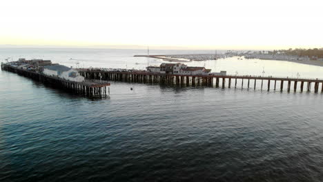 Aerial-drone-shot-flying-above-Stearns-Wharf-pier-at-sunset-with-sailboats-in-the-harbor-of-Santa-Barbara,-California