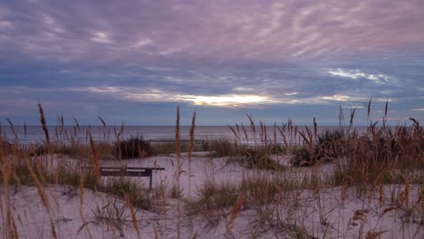 Beautiful-timelapse-of-fast-moving-clouds-over-the-Baltic-sea-coastline,-sunset,-nature-landscape-in-motion,-white-sand-dunes-with-dry-grass-in-foreground,-empty-bench,-wide-angle-shot