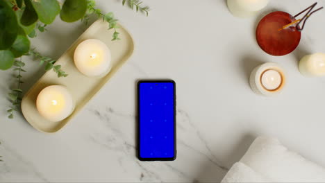 overhead view looking down on still life of blue screen mobile phone lit candles and incense stick with green plant and towels as part of spa day decor 3