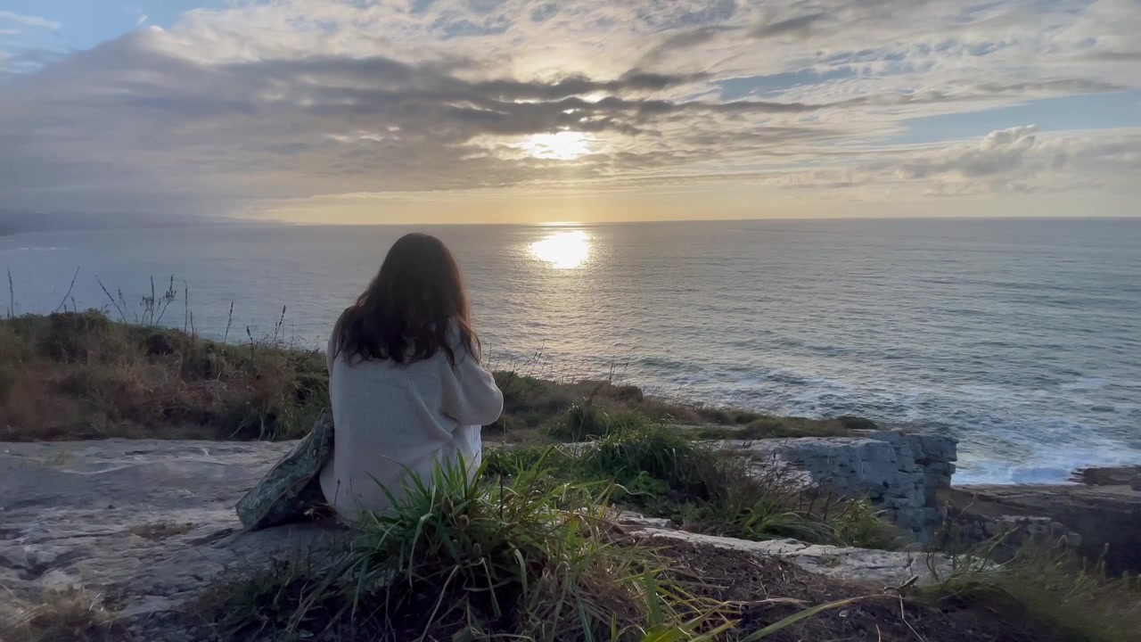 Mujer sentada en un acantilado costero observando el atardecer sobre el mar