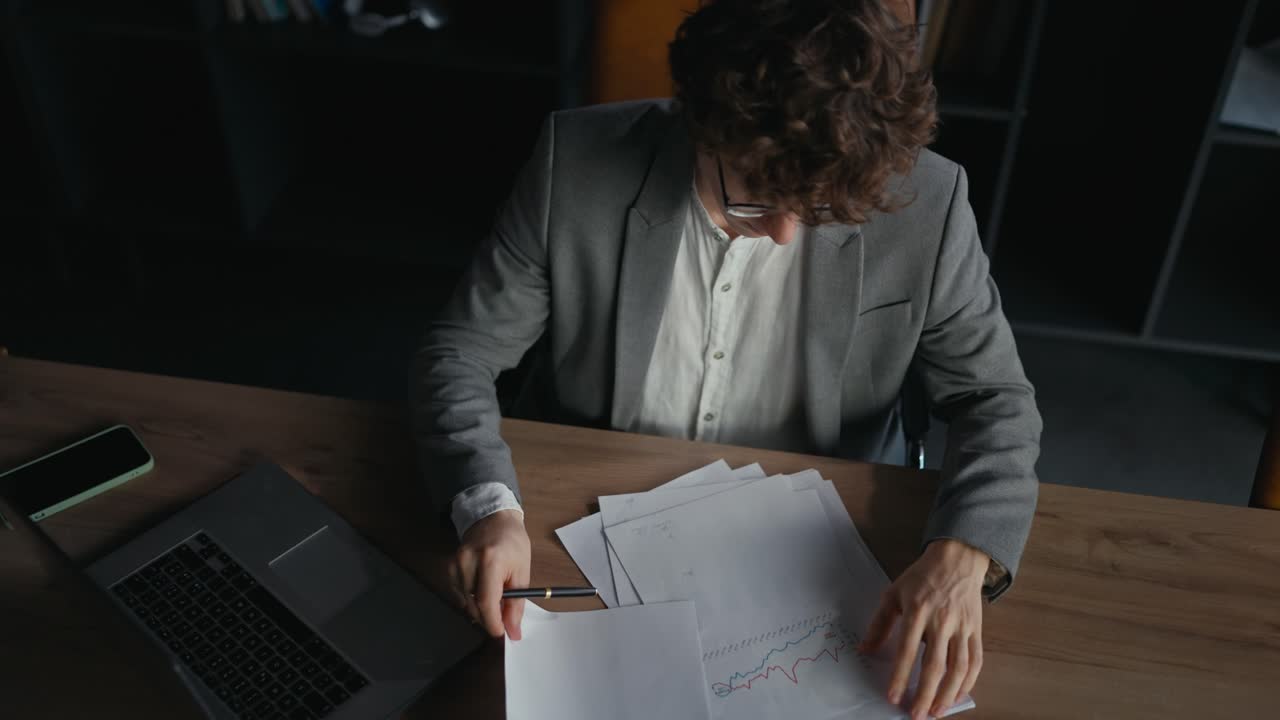 Vista dall'alto di un giovane uomo sicuro di sé con una giacca grigia che esamina grafici, documenti e rapporti di lavoro mentre è seduto a un tavolo in ufficio.