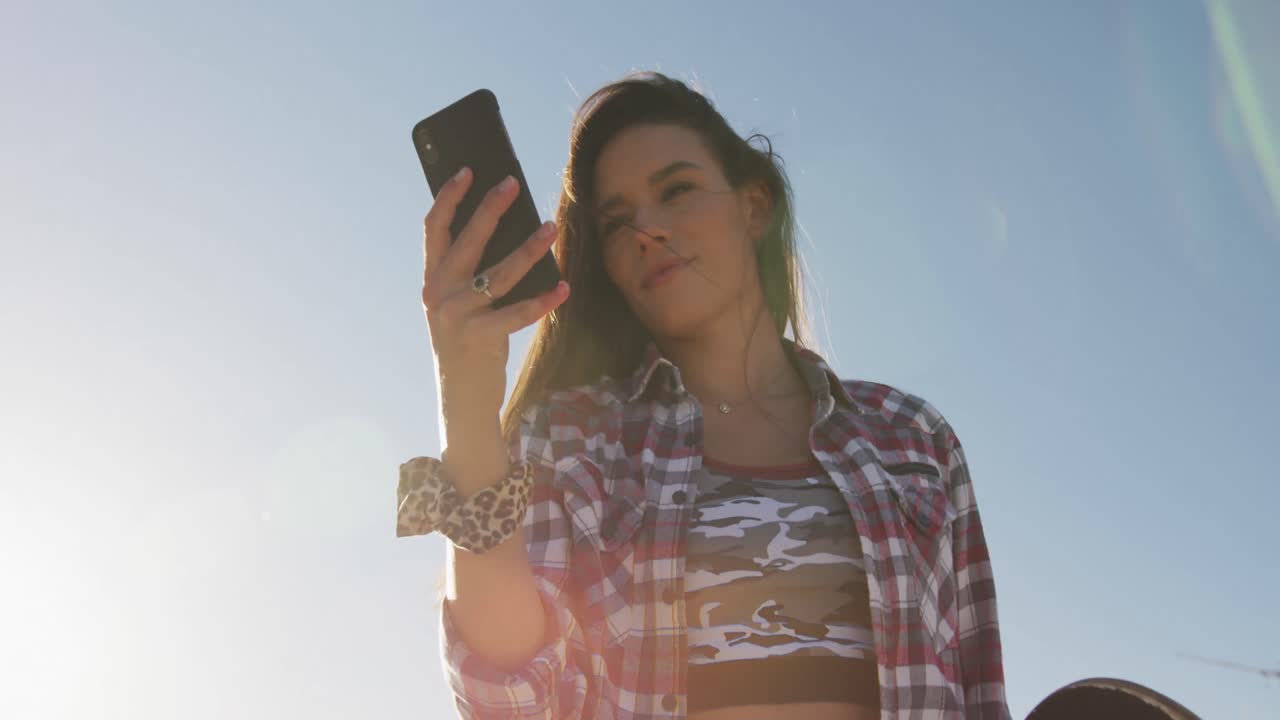 mujer caucásica sonriente usando un teléfono inteligente y sosteniendo un skateboard en un skatepark