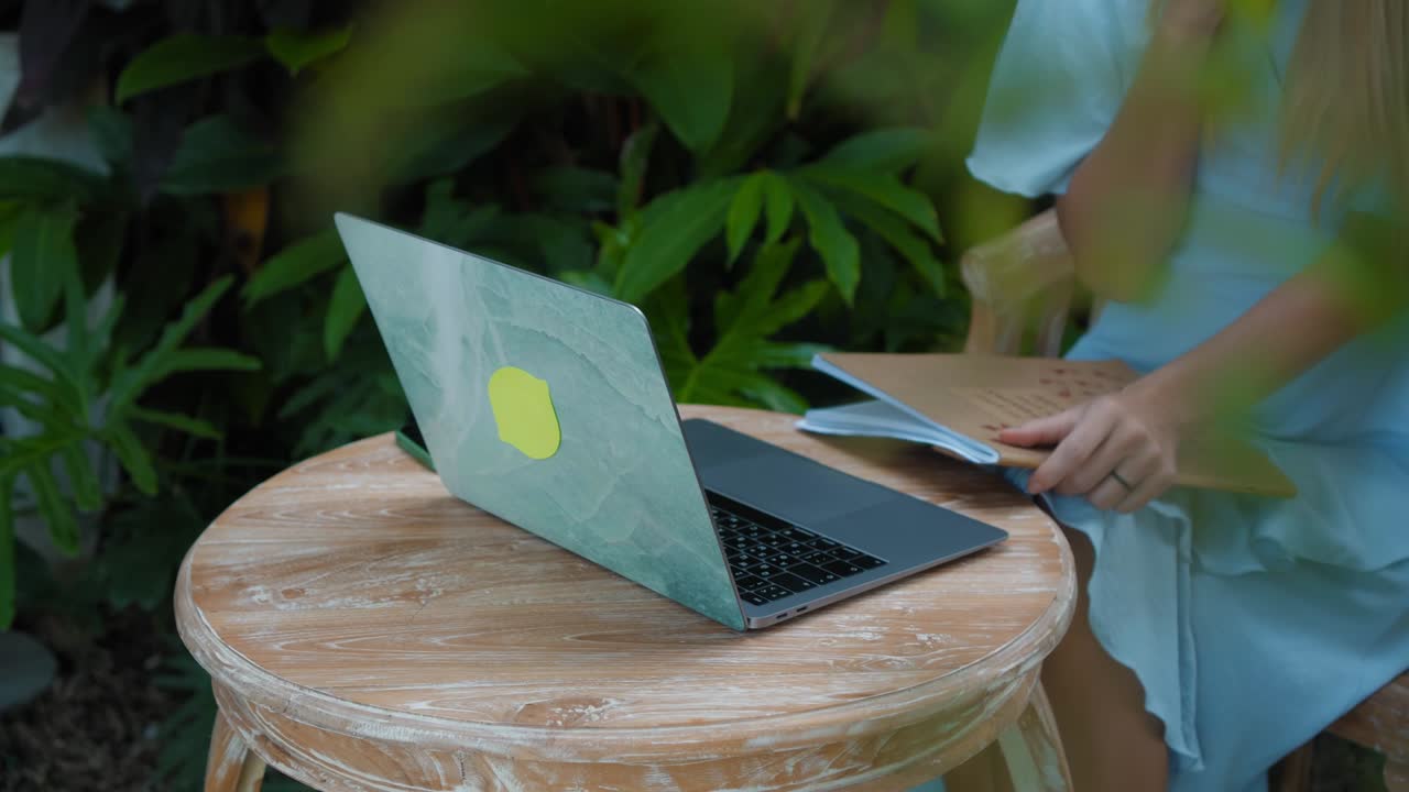 una chica en un vestido azul remotamente en línea trabajando detrás de una computadora portátil y mirando a la pantalla el patio trasero con plantas verdes