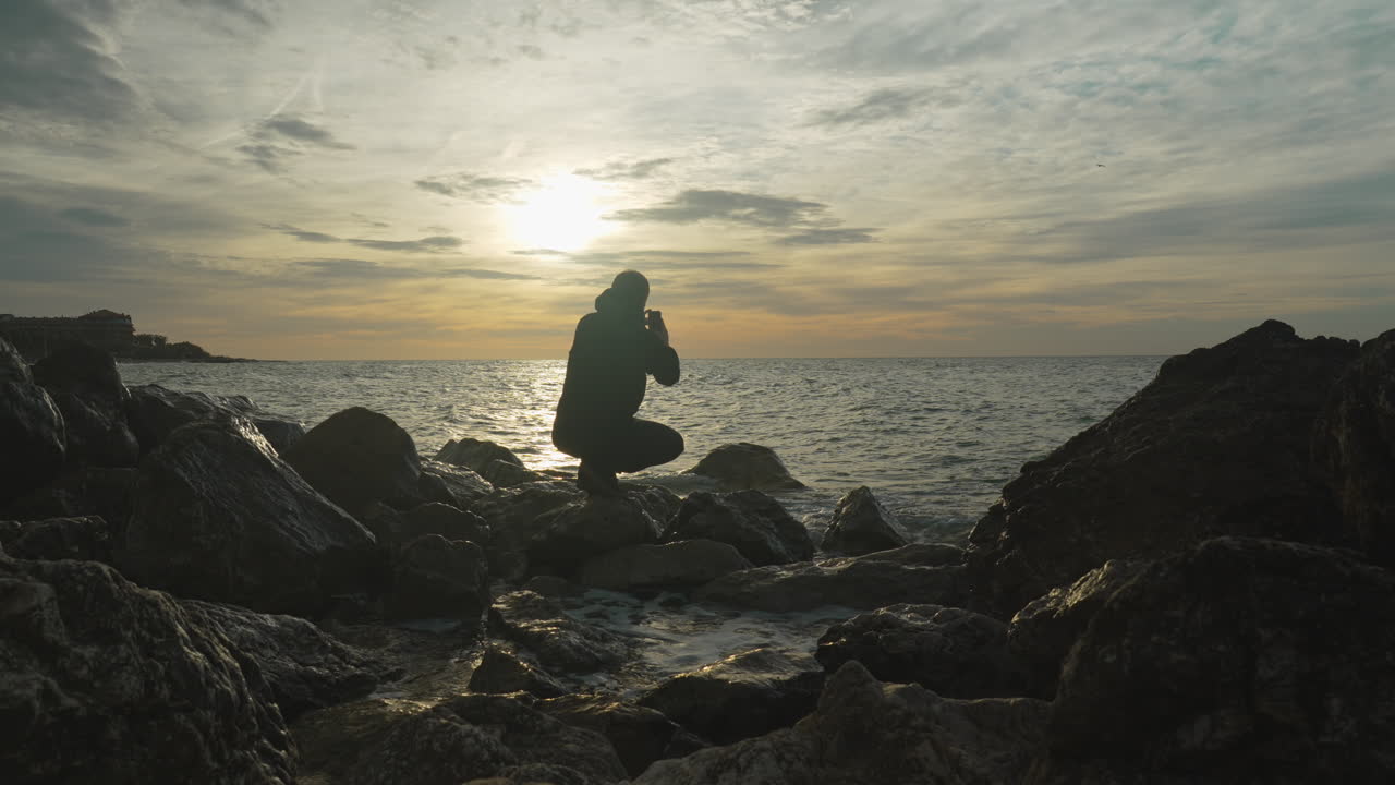 el hombre se sienta en una roca para tomar una foto de la espectacular vista al mar de la hora dorada mientras escapa de las olas que se lavan en tierra