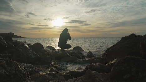 man-sits-down-on-a-rock-to-take-a-photo-of-dramatic-sea-view-of-golden-hour-as-he-escapes-waves-washing-onshore