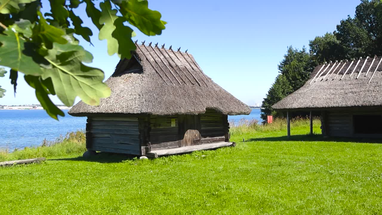 Two traditional old and vintage fishing village log cabins with thatched reed roofs during a sunny summer day by ocean blue sea side with oak tree leaves in the foreground. Grassy area at shoreline
