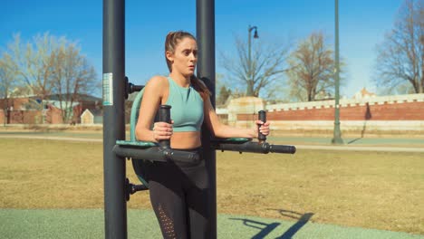 young attractive fitness woman exercising at outdoor gym, doing hanging leg raise exercise while staying focused and determined, wearing sports bra and yoga pants