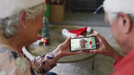 Lächelndes-älteres-Kaukasisches-Paar,-Das-Sein-Smartphone-Für-Einen-Weihnachtsvideoanruf-Mit-Einem-Mann-Auf-Dem-Bildschirm-Nutzt